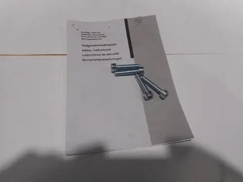 Blatt mit Montageanleitung von Remeha und mehreren Schrauben. Die Anleitung enthält Sicherheitshinweise in mehreren Sprachen, darunter Englisch und Deutsch.